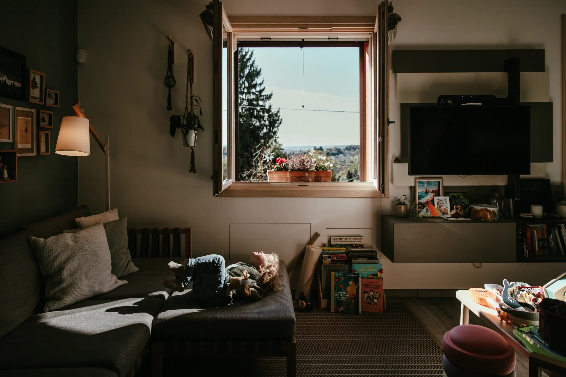 Bambino disteso sul divano in un soggiorno luminoso con vista dalla finestra, fotografia di famiglia in interni scattata da Sara Mellerio.
