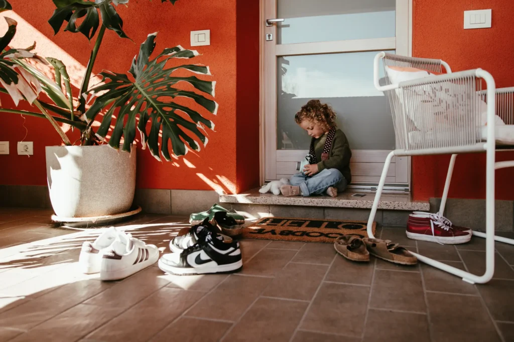 famiglia-03-26-2 Bambino che gioca con una macchina fotografica giocattolo sulla soglia di casa con piante e scarpe, servizio fotografico di famiglia all'aperto di Sara Mellerio.