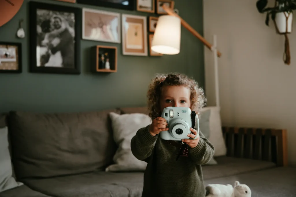 famiglia-03-26-3 Primo piano di un bambino che tiene una fotocamera istantanea azzurra davanti al viso, ritratto creativo di famiglia di Sara Mellerio.