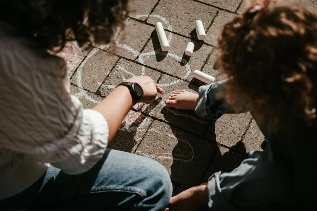 famiglia-03-26-5 Dettaglio artistico di mani e piedi di un bambino che gioca con gessetti colorati sul pavimento esterno, scatto di famiglia di Sara Mellerio.