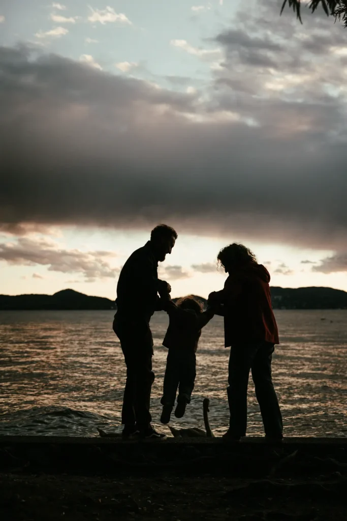 famiglia-03-26-7 Genitori che fanno dondolare il bambino per mano davanti a un lago al crepuscolo, servizio fotografico di famiglia emozionale di Sara Mellerio.