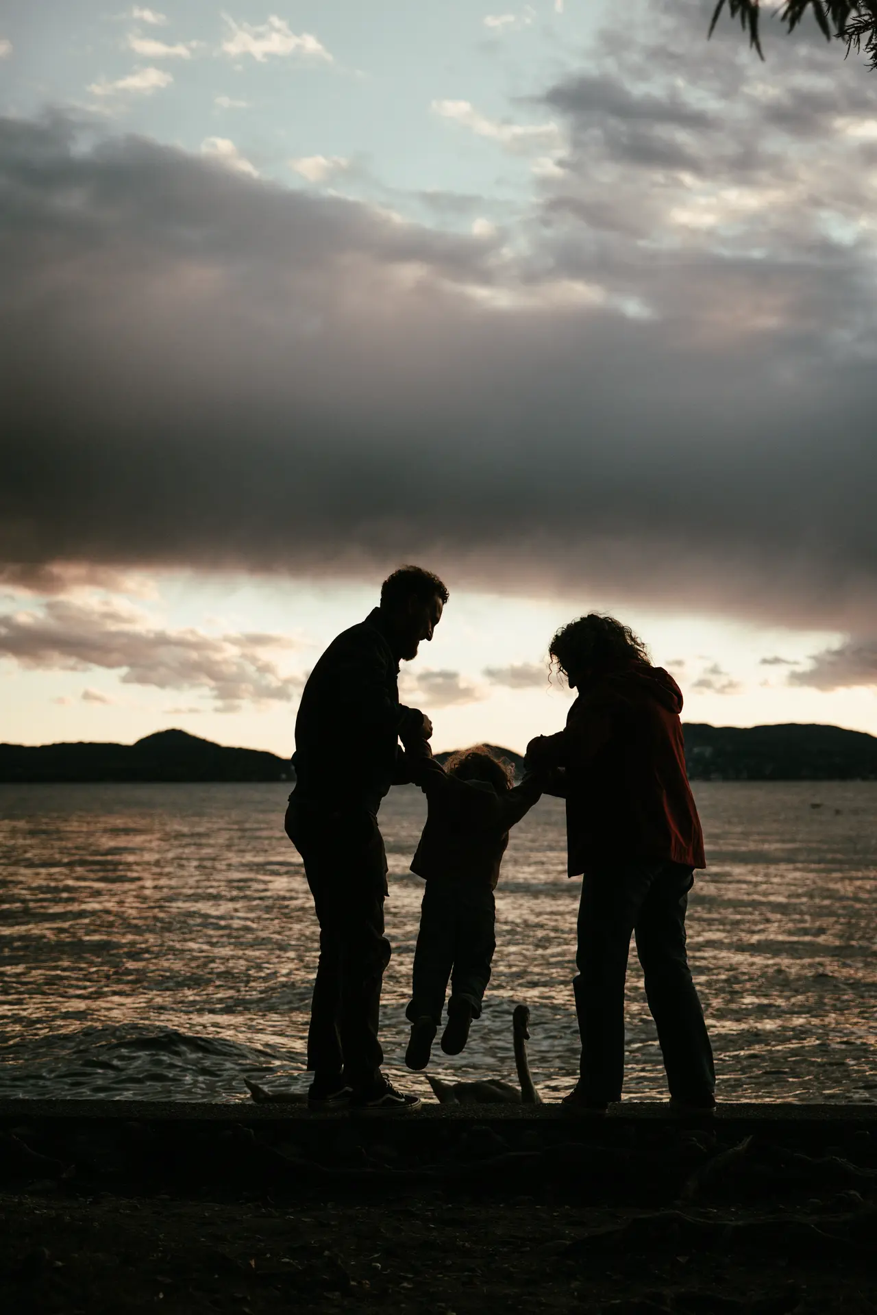 Genitori che fanno dondolare il bambino per mano davanti a un lago al crepuscolo, servizio fotografico di famiglia emozionale di Sara Mellerio.