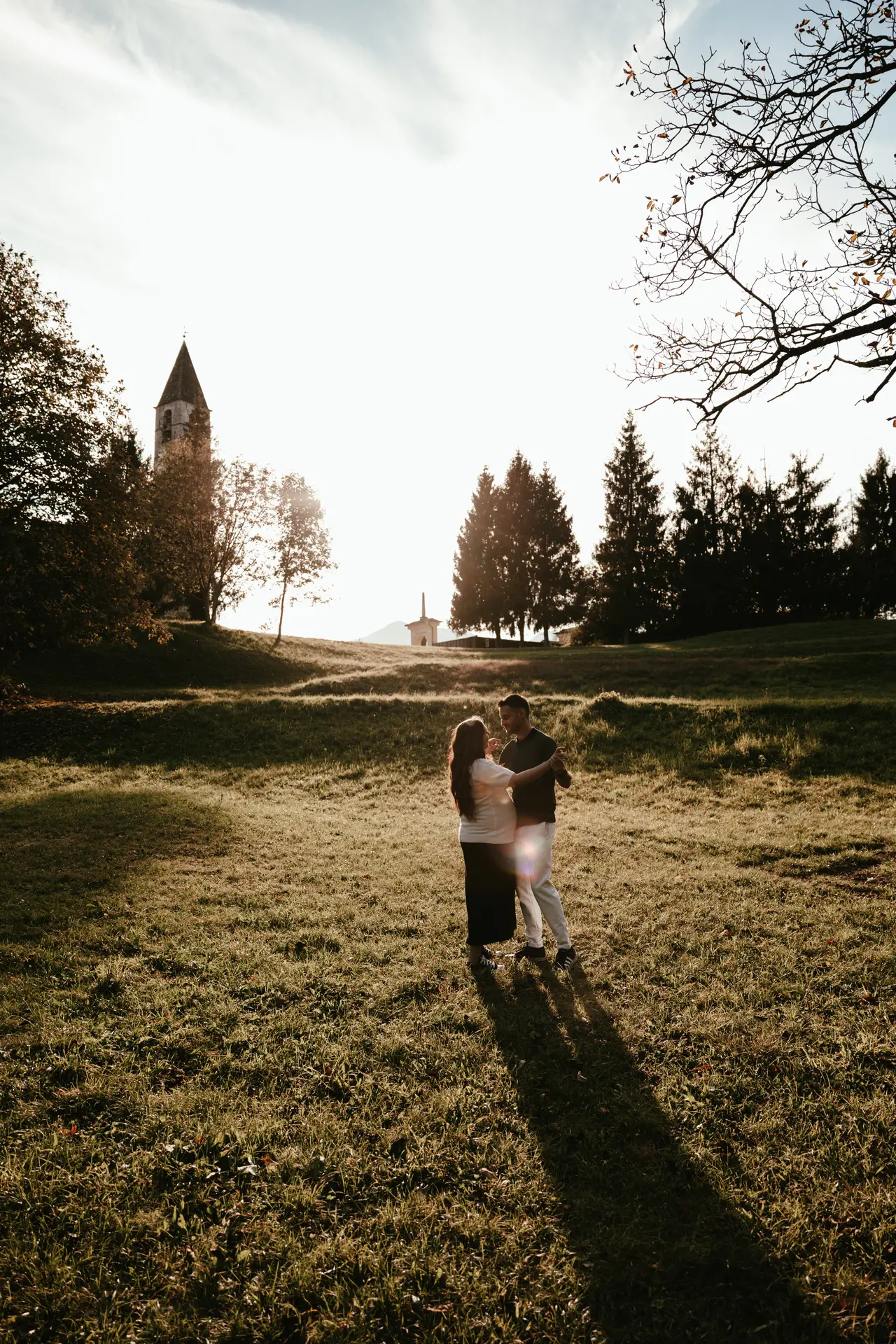 Futuri genitori che ballano in un prato al tramonto con un campanile sullo sfondo, scatto premaman di Sara Mellerio.