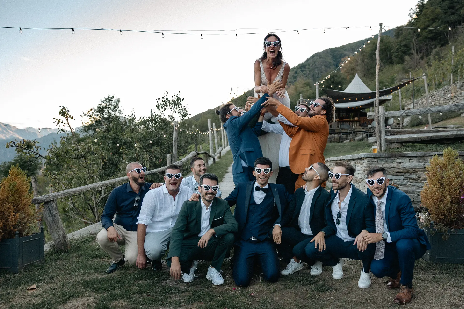 Gli amici dello sposo sollevano la sposa indossando occhiali a cuore divertenti, fotografia creativa di Sara Mellerio.
