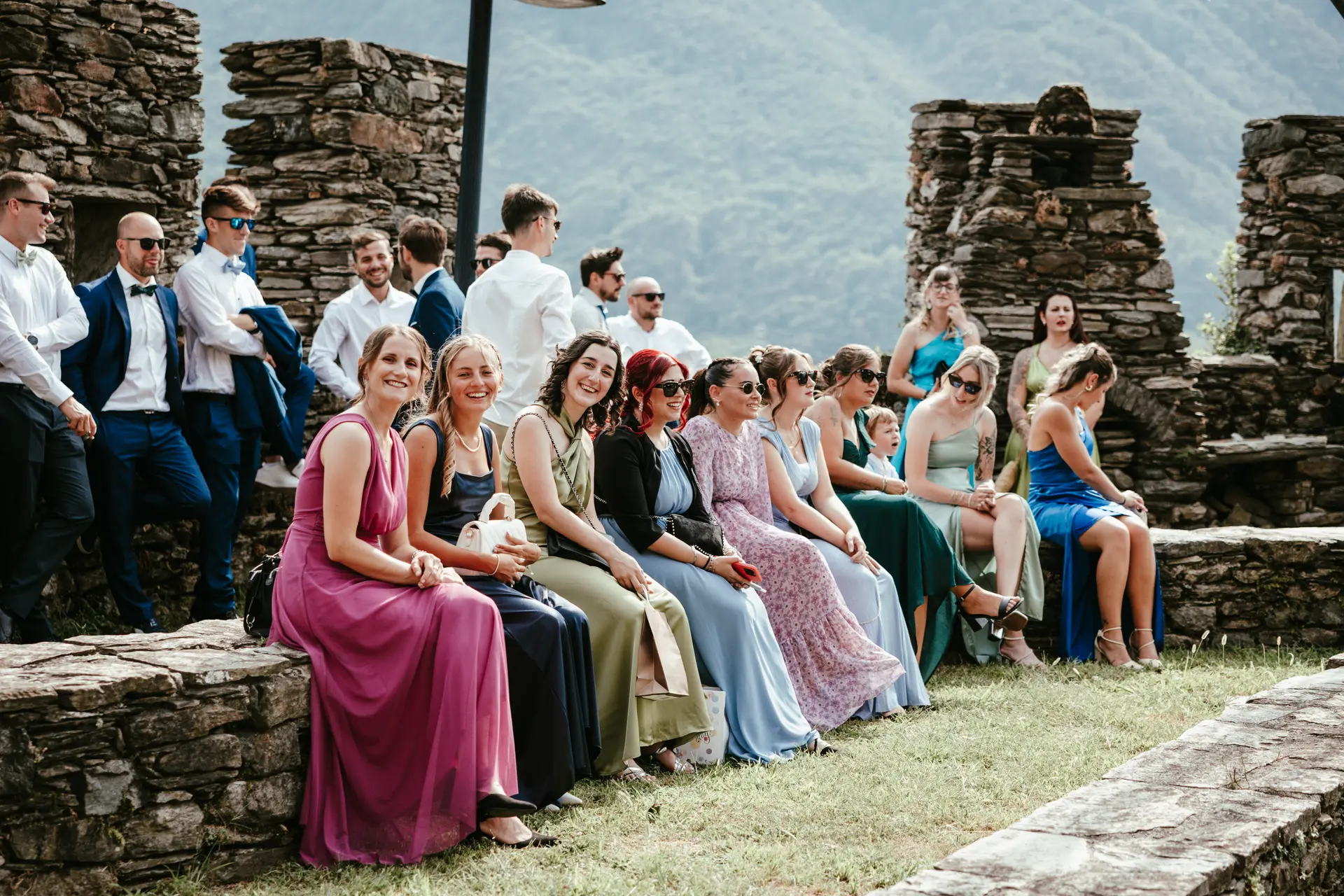 Gruppo di invitate al matrimonio sedute su un muretto in pietra in una location panoramica, fotografia di reportage di Sara Mellerio.
