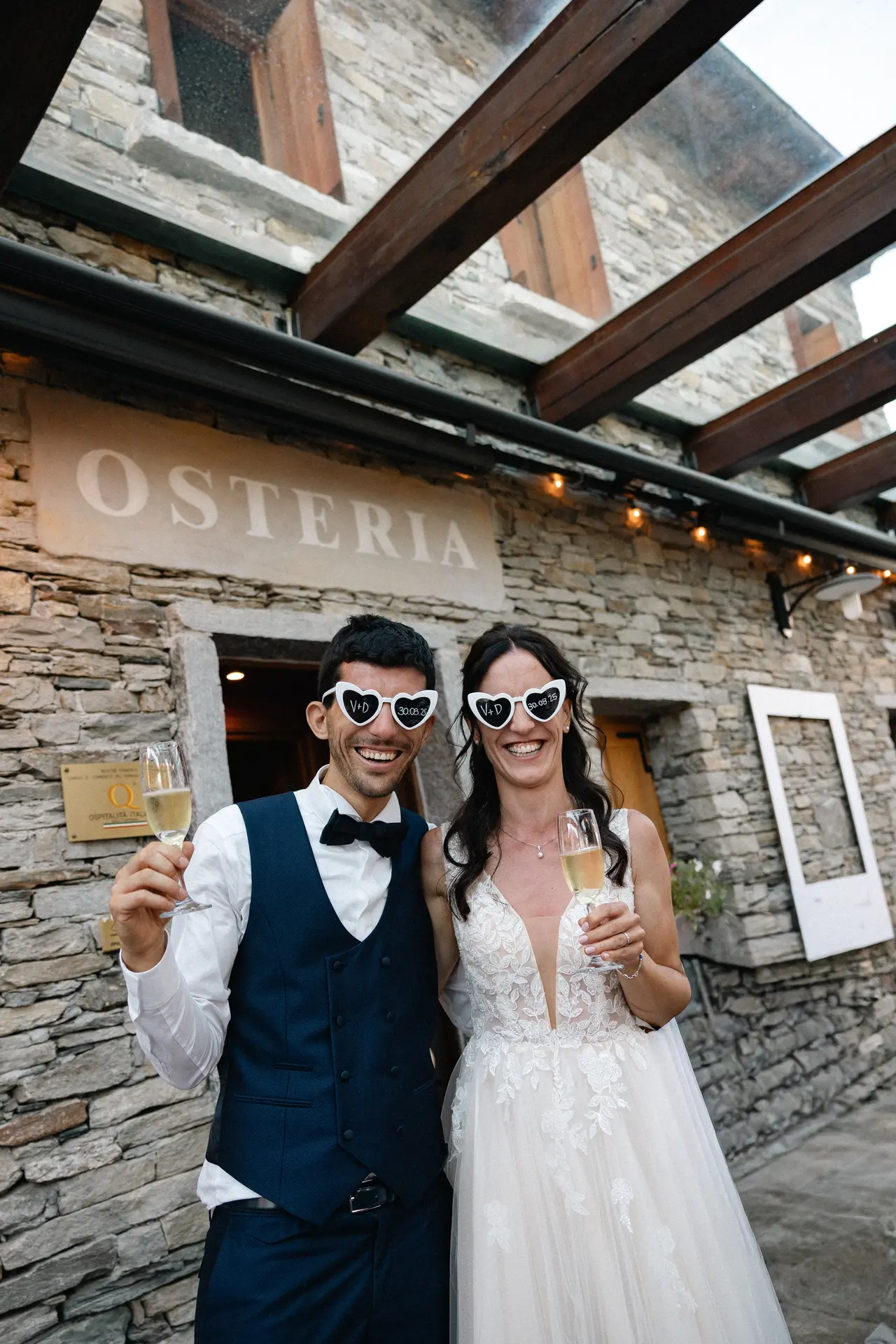 Ritratto degli sposi con occhiali a cuore e calici di spumante davanti a un'osteria storica, fotografia di Sara Mellerio.